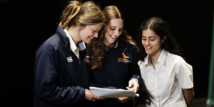 Three female students at Rototuna High School smiling and reviewing a document together in a classroom setting.